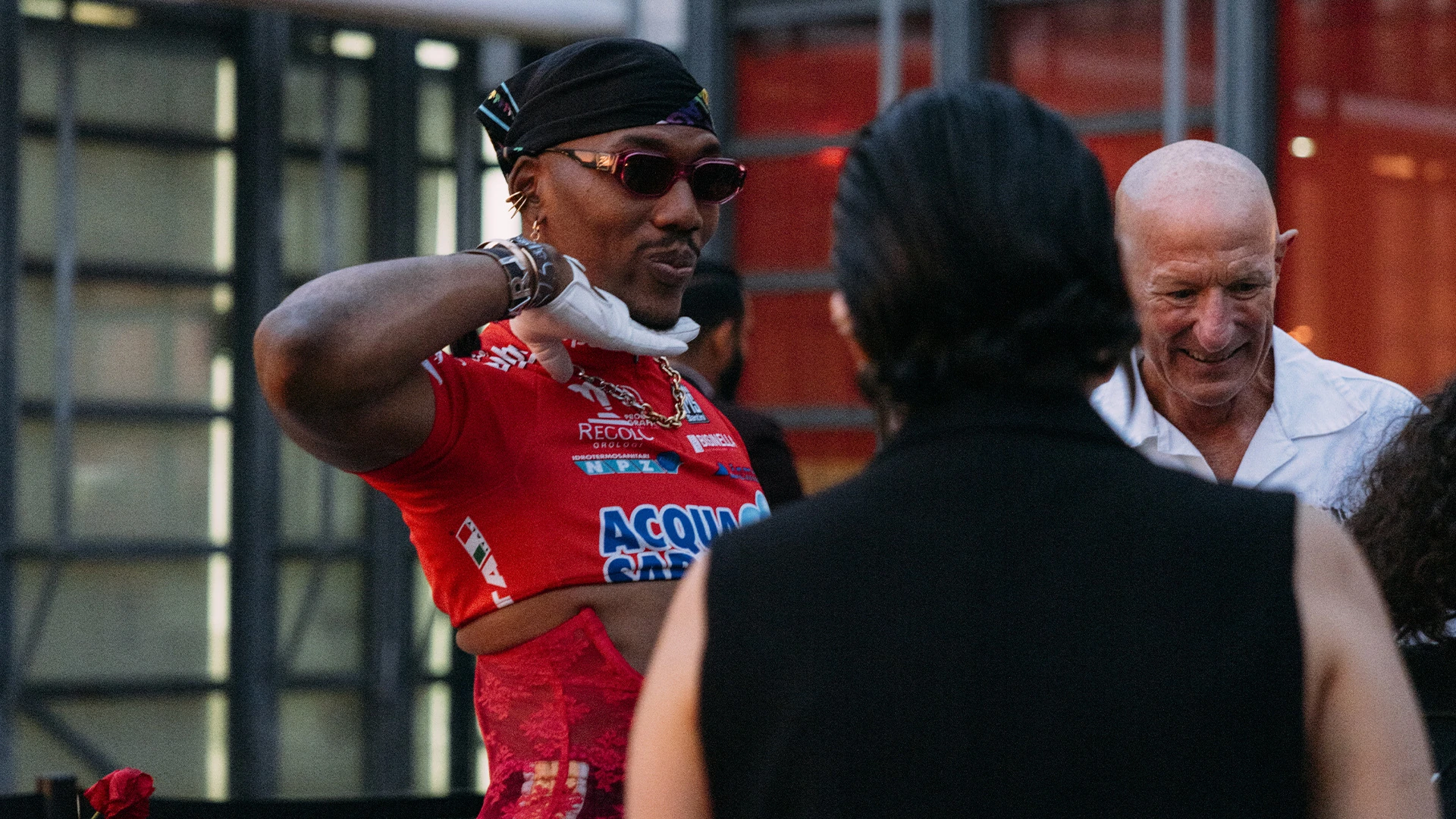 A guest in a red racing jersey and sunglasses talking on a rooftop terrace.