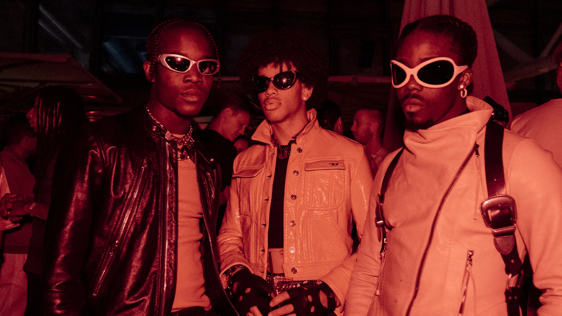 Three guests in wraparound sunglasses and leather jackets under warm red light.