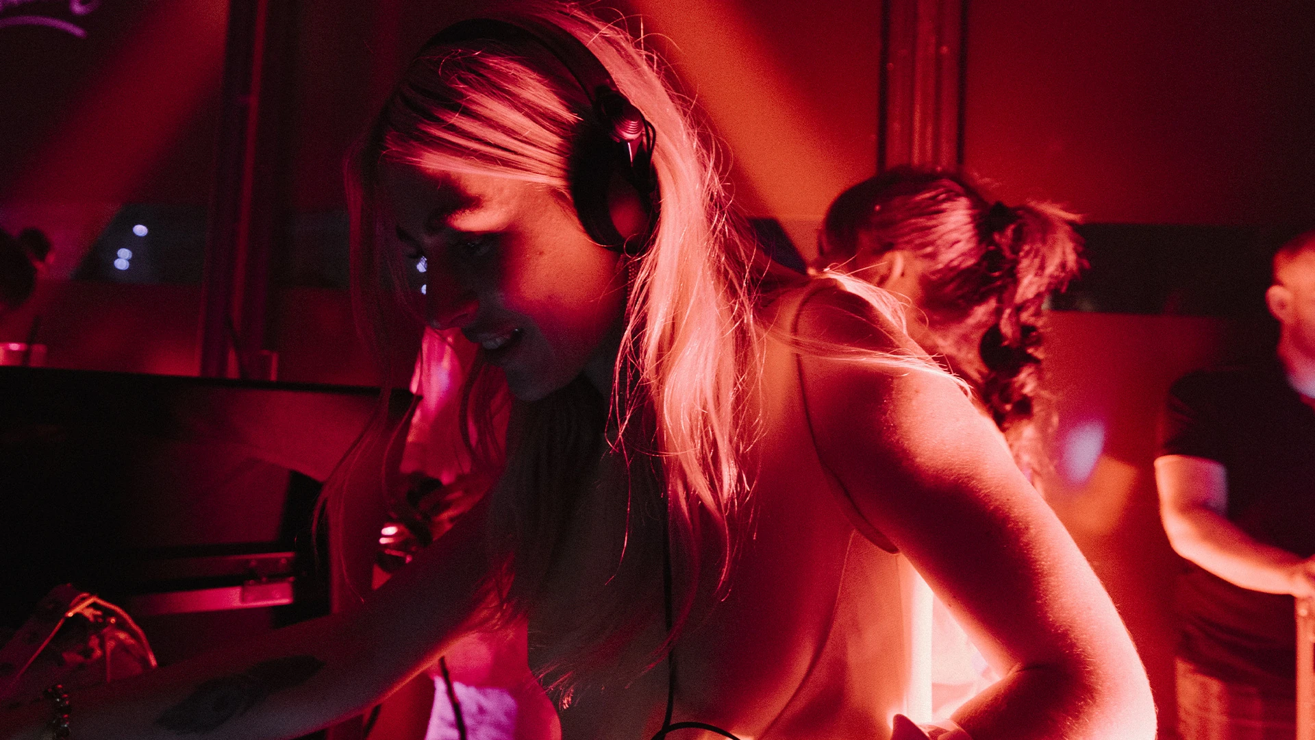 A DJ in headphones leaning over the decks under deep red light.