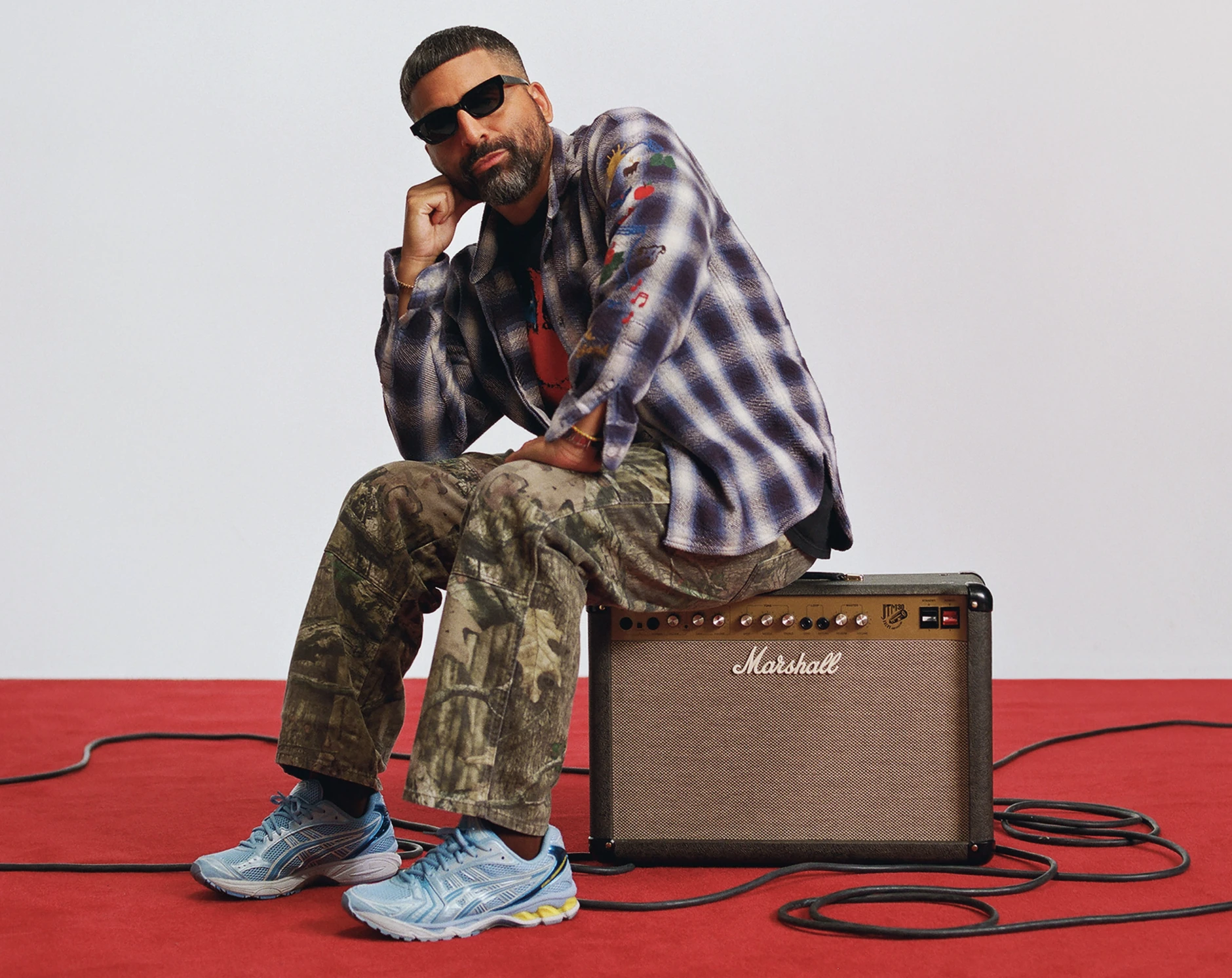 Angelo Baque perched on a Marshall combo amplifier, cables running across a red floor.