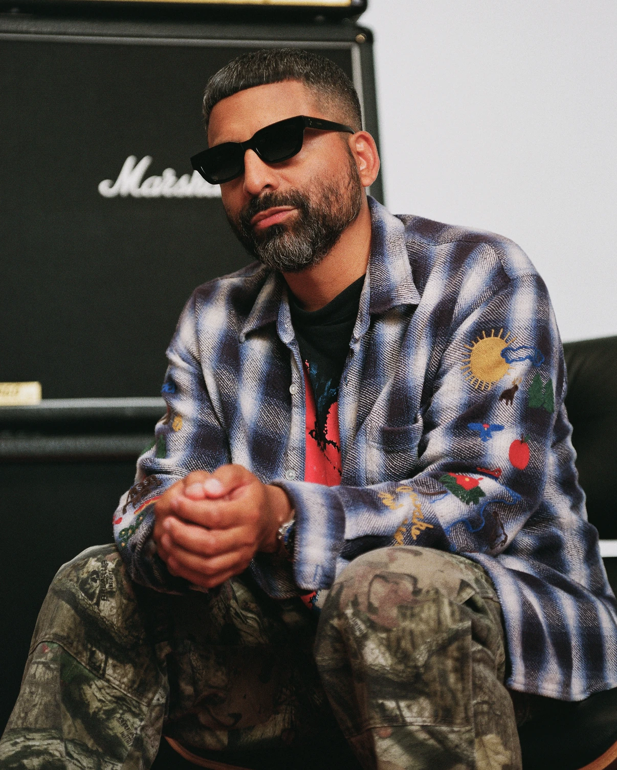 Close portrait of Angelo Baque in sunglasses and an embroidered plaid shirt in front of a Marshall amplifier.