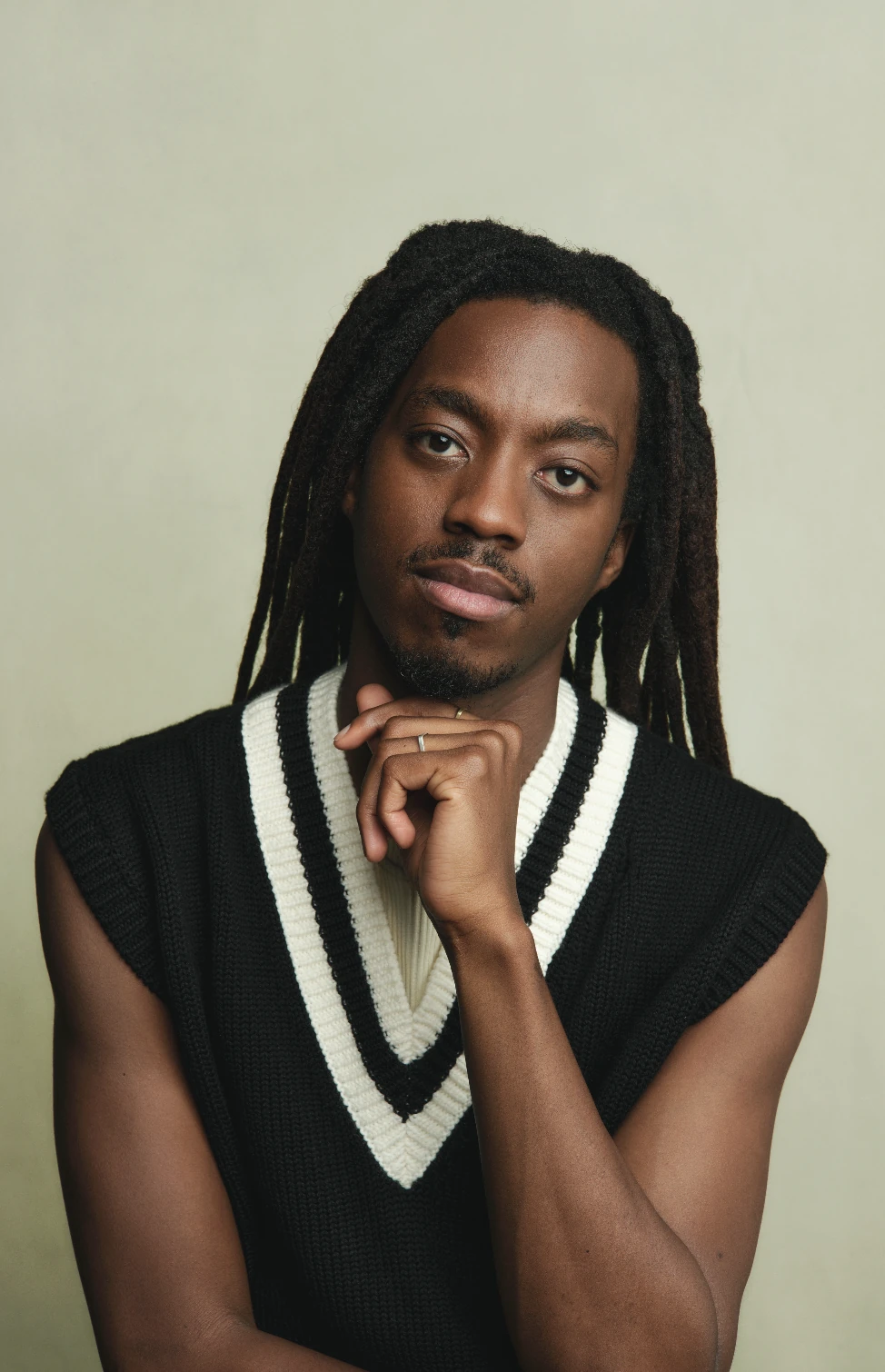 Studio portrait of a model with locs in a black and white V-neck vest, hand to chin.