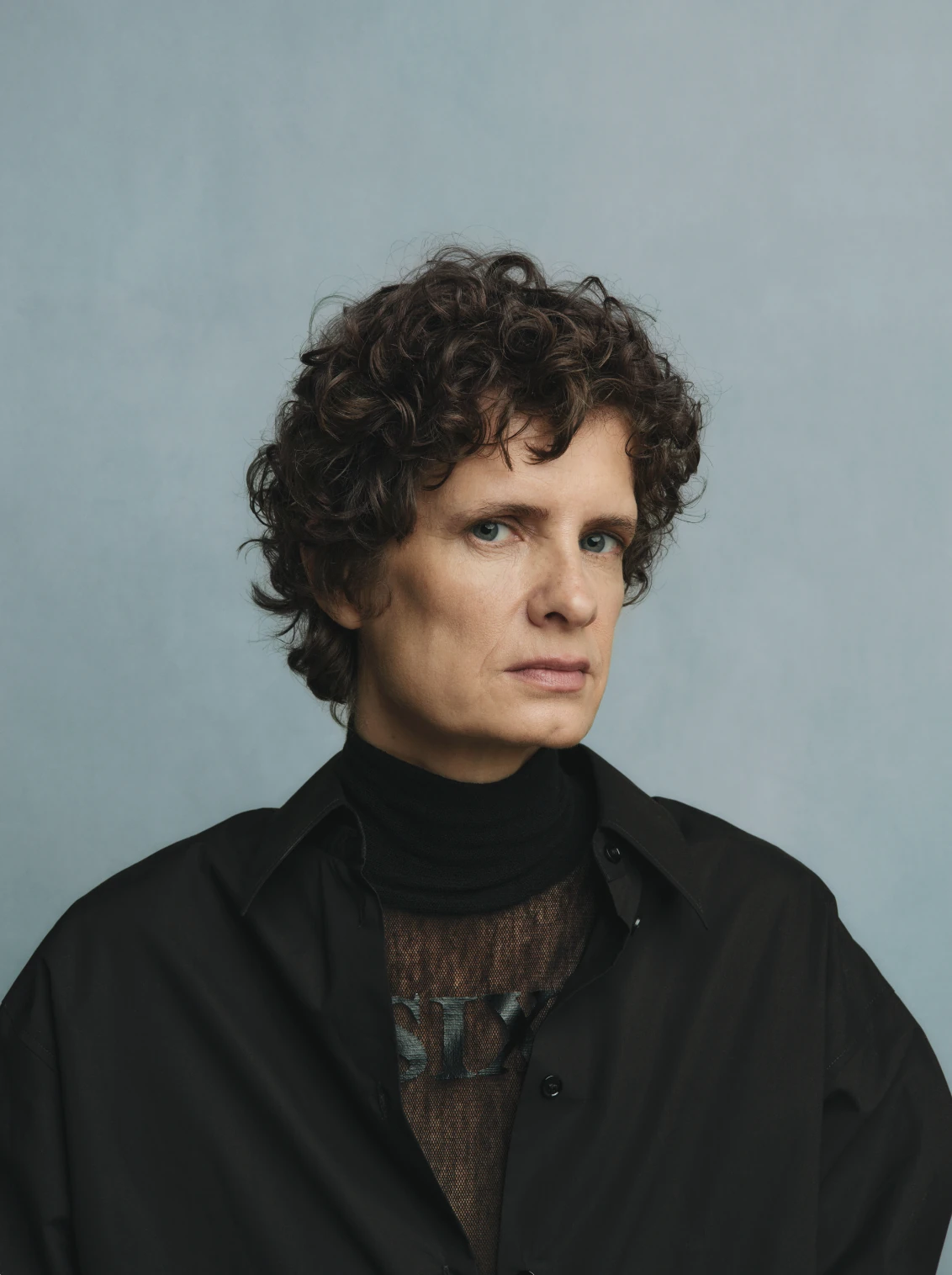 Studio portrait of a model in a black overcoat and turtleneck against a pale blue backdrop.