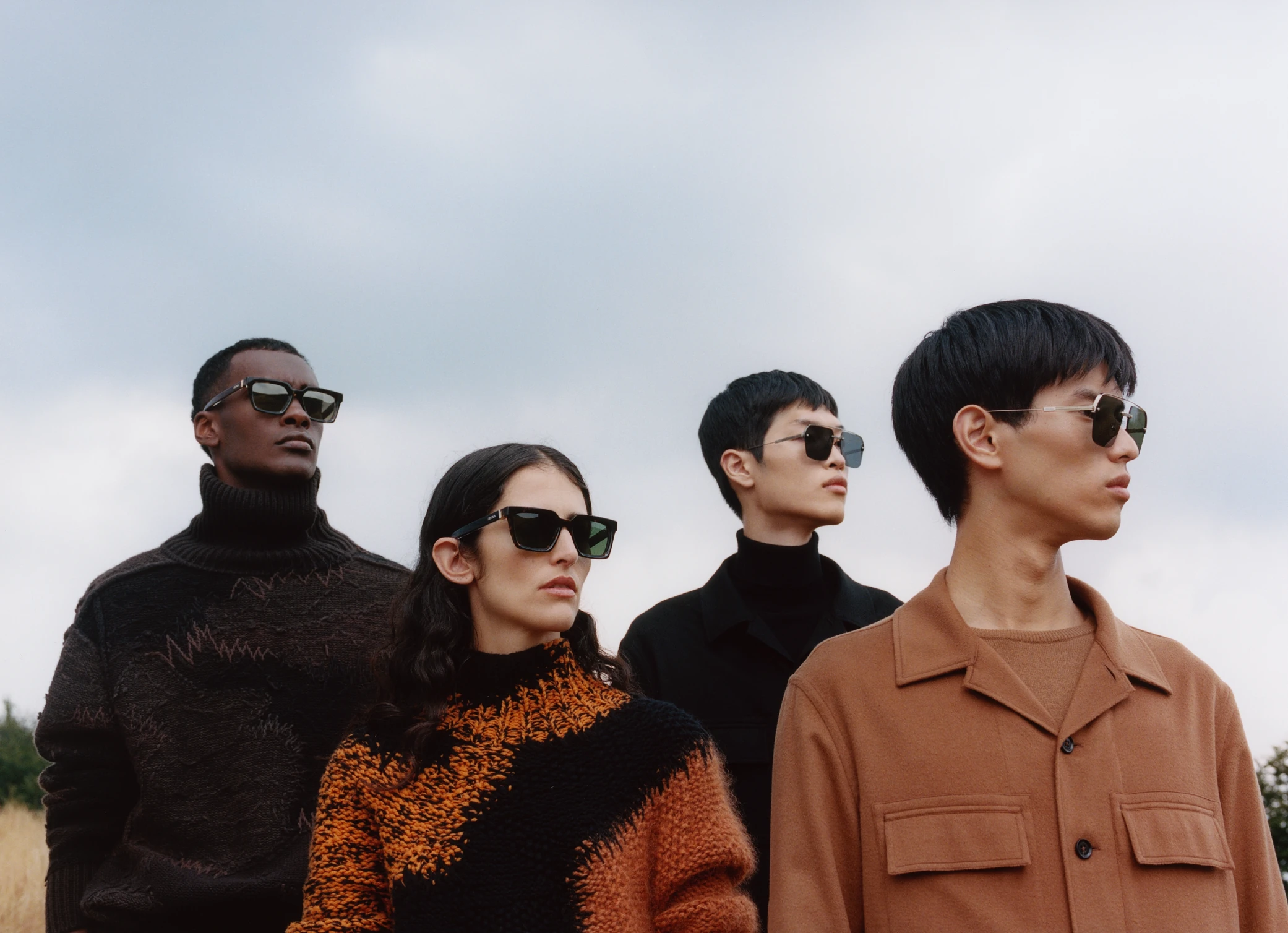 Four models in sunglasses and autumn-toned knitwear grouped against an overcast sky.