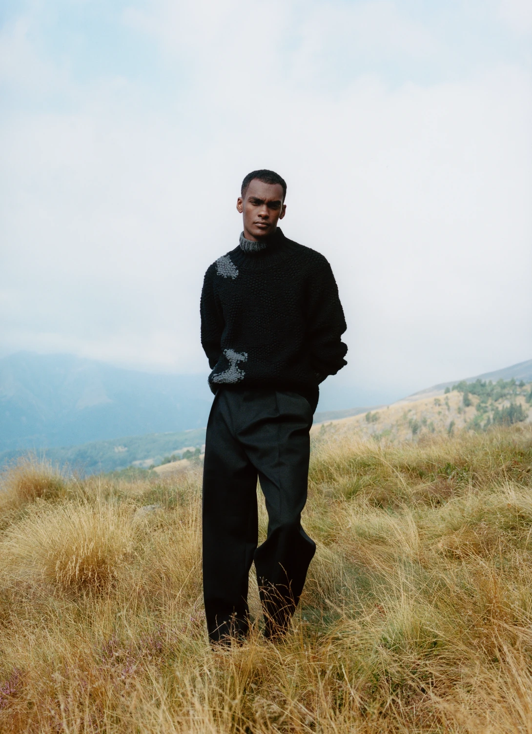 A model in a black cashmere sweater and wide black trousers on a grassy ridge above hazy mountains.