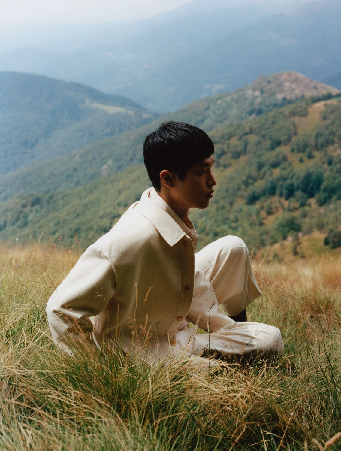 A model in a cream overshirt and trousers sitting in hillside grass, looking out over forested valleys.