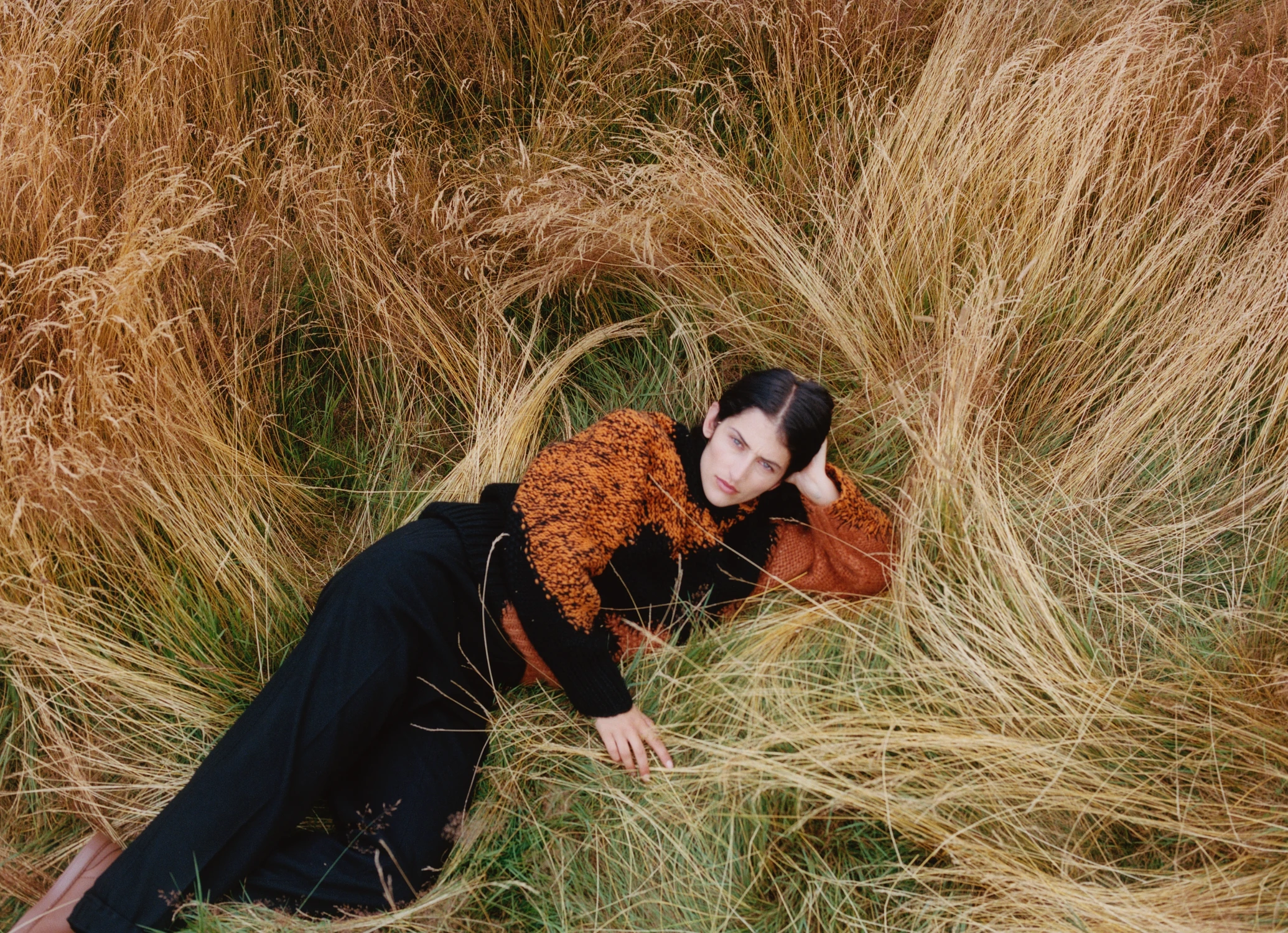 A model lying in tall golden grass in a black coat and textured orange cashmere top, shot from above.