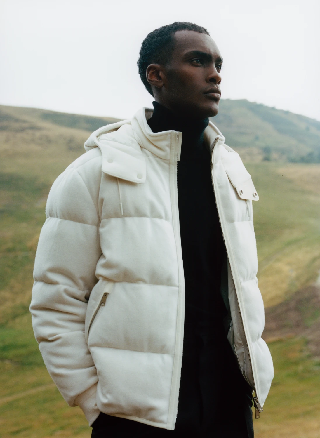 A model in a cream padded overshirt and black turtleneck standing against green hills under an overcast sky.