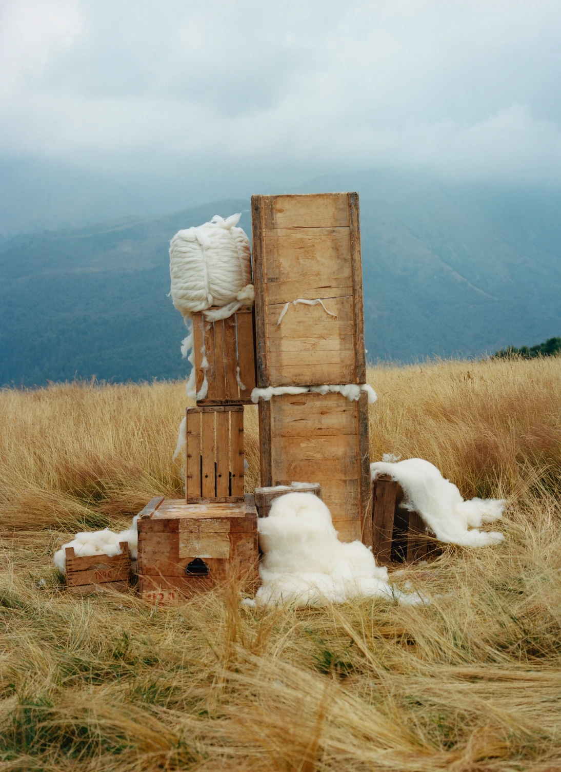 Stacked wooden crates and raw white cashmere fibre arranged in a windswept hilltop meadow.