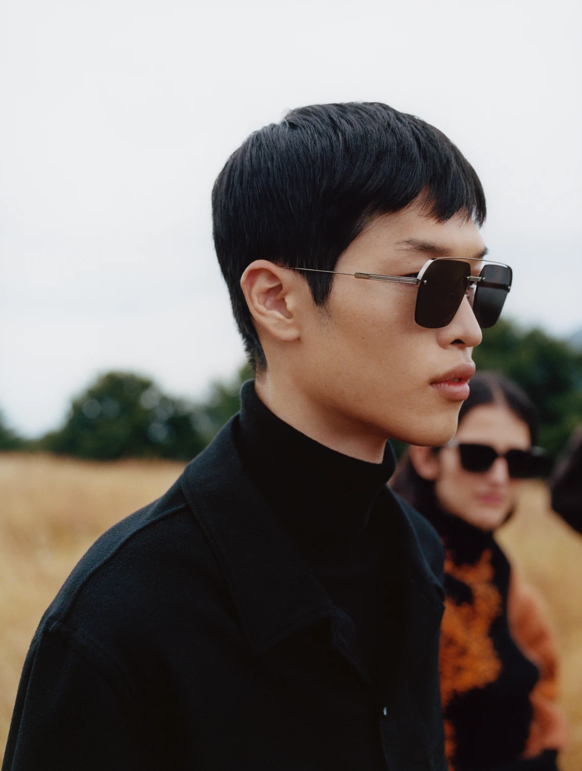 Close portrait of a model in dark sunglasses and a black turtleneck, a second model in orange knitwear behind.