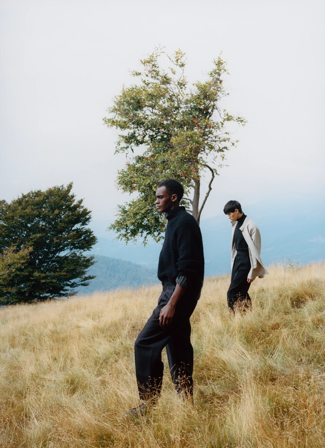 Two models walking through a dry hillside meadow beside a lone tree, one in black knitwear, one in a pale coat.