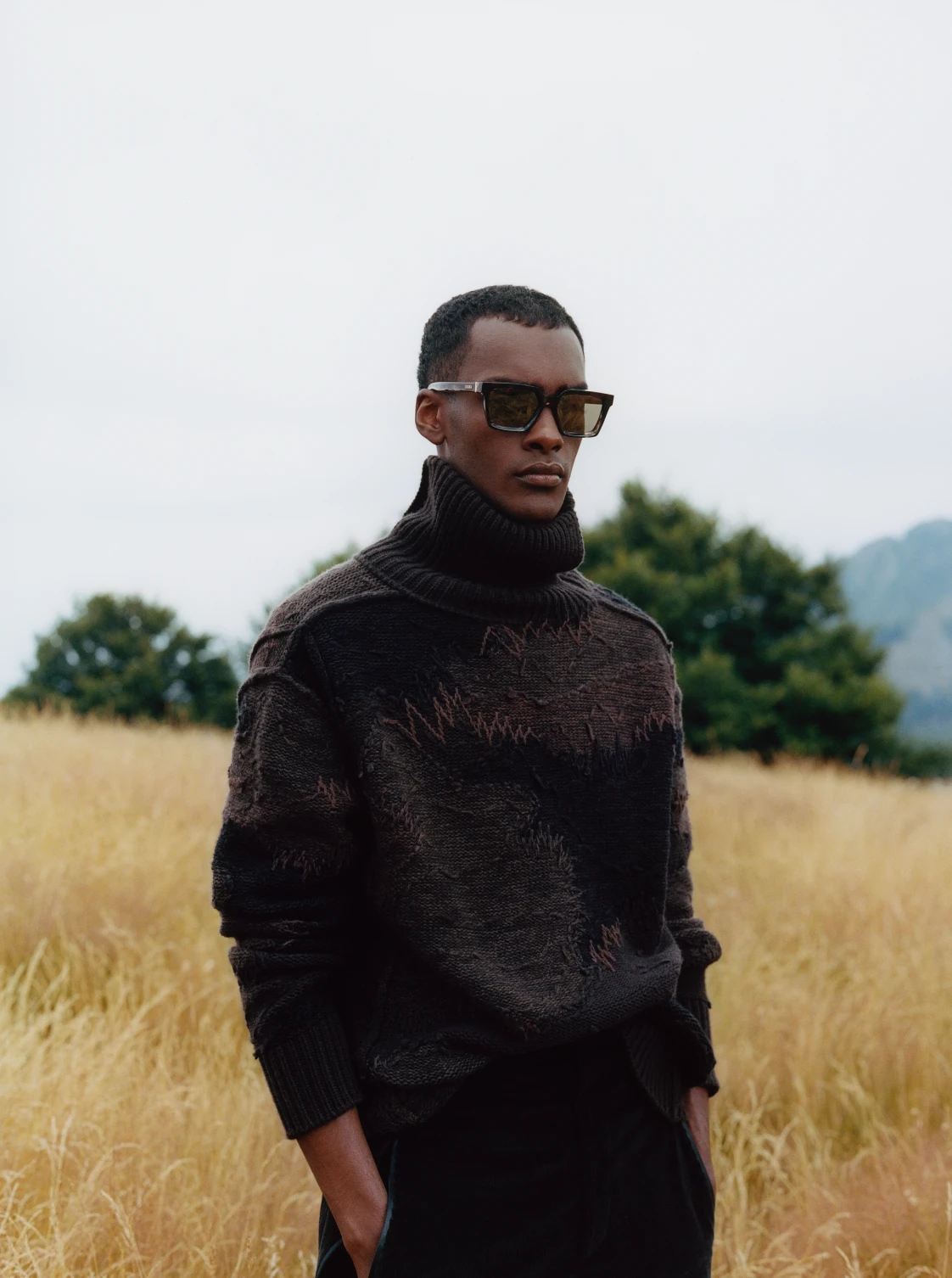 A model in dark sunglasses and a charcoal patterned cashmere turtleneck standing in a field of pale grass.